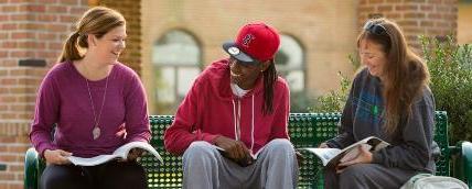 Three students sitting on an outdoor bench on campus. They are laughing and holding open textbooks.